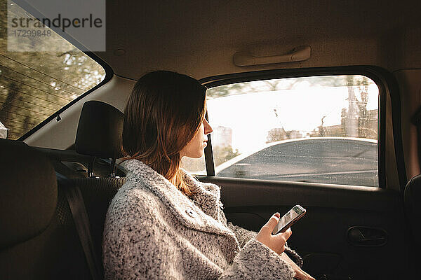Frau schaut durch das Fenster und hält ihr Smartphone im Auto
