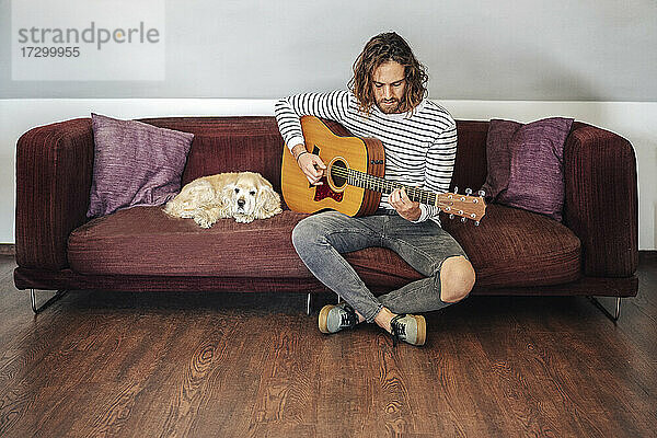 Attraktiver Mann mit langen Haaren spielt akustische Gitarre im Haus mit Hund