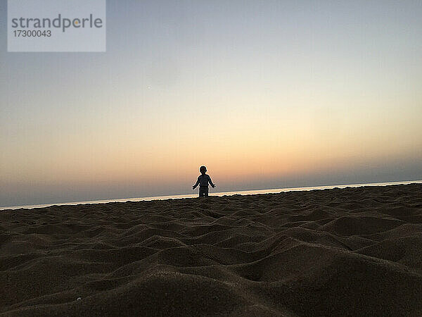 Ein kleines Kind steht am Strand und streckt bei Sonnenuntergang die Hände aus.