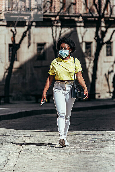 Schwarzes afroamerikanisches Mädchen mit Gesichtsmaske  das eine Stadtstraße entlanggeht.