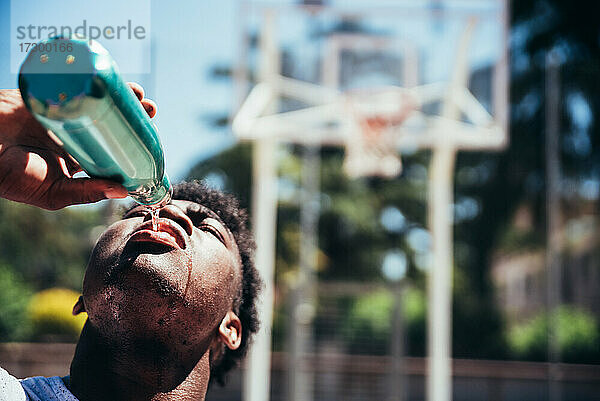 Porträt eines afroamerikanischen schwarzen Jungen  der nach dem Training auf einem städtischen Basketballplatz Wasser trinkt.