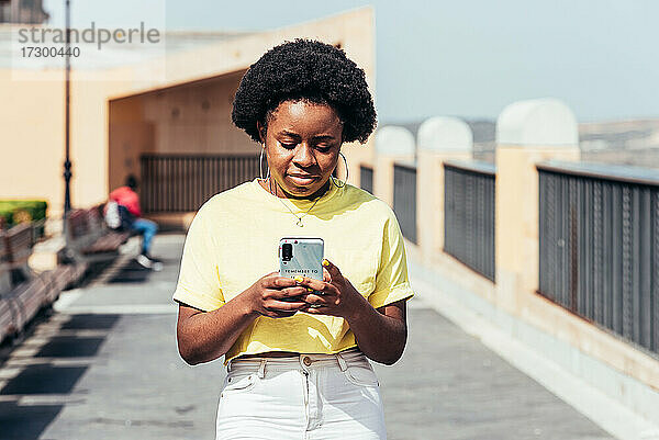 Porträt eines schwarzen Mädchens mit Afro-Haar und Ohrringen  das ihr Handy benutzt und in einem städtischen Raum spazieren geht.