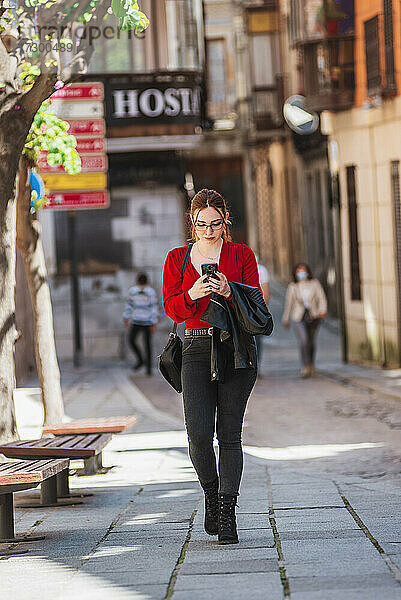 Junges rothaariges Mädchen mit Brille  roter Bluse und Jeans  das eine Straße in der Stadt entlangläuft und dabei ihr Mobiltelefon benutzt