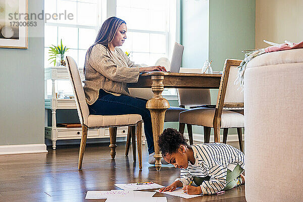 Niedliches Mädchen  das auf Papier zeichnet  während die Mutter einen Laptop benutzt  während sie am Tisch im Wohnzimmer sitzt