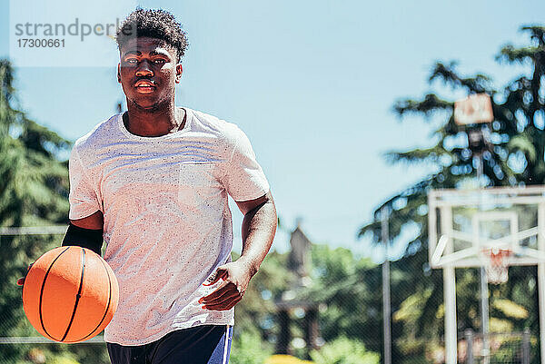 Porträt eines afro-amerikanischen schwarzen Jungen  der auf einem städtischen Platz Basketball spielt. Er läuft und wählt den Ball.