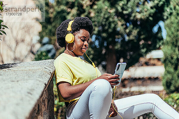 Schwarzes afroamerikanisches Mädchen sitzt und entspannt  während sie mit ihrem Handy und Kopfhörern Musik hört.