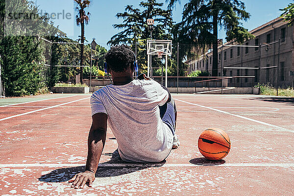 Rückansicht eines schwarzen Afro-Jungen  der sein Mobiltelefon benutzt und mit Kopfhörern auf dem Basketballplatz Musik hört. Konzept Technologie und Sport.
