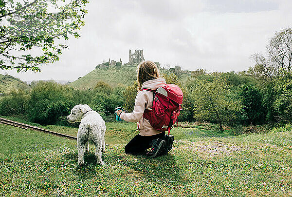 mädchen sitzt mit ihrem hund und schaut auf eine burg in der englischen landschaft