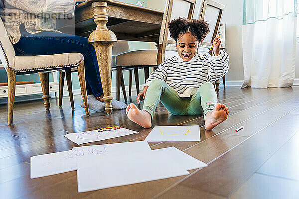 Glückliches Mädchen  das auf Papieren zeichnet  während es zu Hause auf dem Hartholzboden sitzt