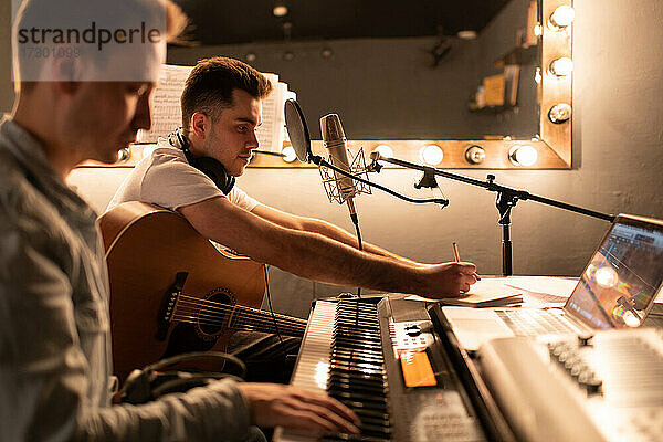 Männlicher Gitarrist macht sich Notizen  während er mit einem Freund in einem modernen Studio einen Song schreibt