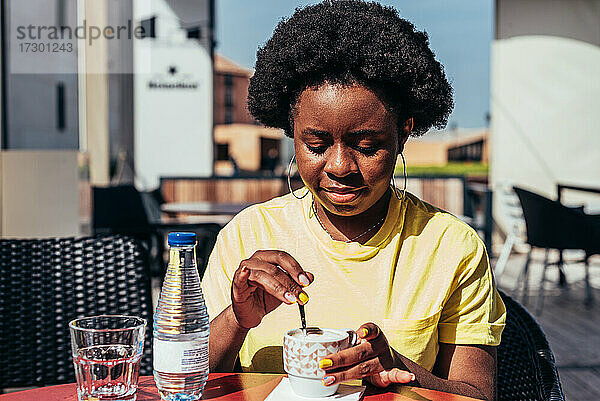Porträt eines schwarzen Mädchens mit Afro-Haar und Ohrringen  das Kaffee und eine Flasche Wasser auf einer Bar-Terrasse trinkt.