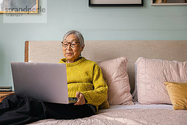 Ältere asiatische Frau  die einen Laptop benutzt  während sie im Schlafzimmer zu Hause sitzt