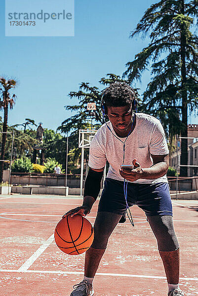 Schwarzer afroamerikanischer Junge  der mit Kopfhörern und seinem Mobiltelefon Musik hört und auf einem städtischen Platz Basketball spielt.