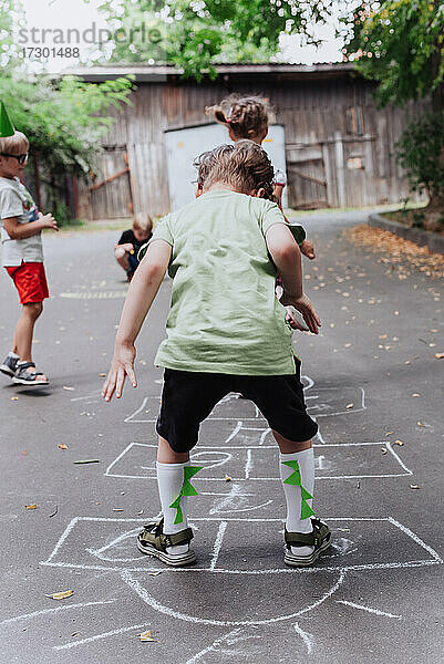 5 Jahre alt Junge Kind spielt hopscotch im Freien  Kinder Outdoor-Aktivitäten  trägt grünes T-Shirt mit Dinosaurus Druck und Dino Helm