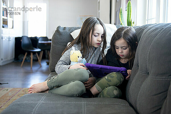 Zwei kleine Mädchen sitzen auf der Couch und benutzen ein Tablet.