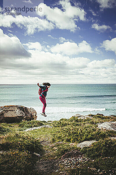 Dunkelhaarige Frau mit rotem Rucksack  die von einem Felsen am Meer springt