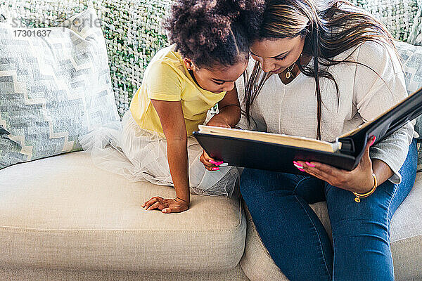 Mutter schaut sich mit ihrer Tochter ein Fotoalbum an  während sie auf dem Sofa im Wohnzimmer sitzt