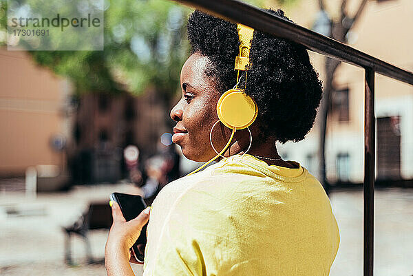 Rückansicht eines schwarzen Mädchens mit Afro-Haar und Ohrringen  das mit ihrem Handy und gelben Kopfhörern in einem städtischen Raum Musik hört.