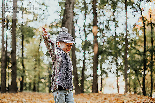 Stilvoller Junge  der im Herbst im Stadtpark Spaß hat. Glückliches Kind spielt mit gefallenen Blättern. Kindermode. Junge trägt trendigen gelben Mantel  Mütze und Schal. Lächelnder kleiner Junge im Freien. Kind springt und rennt