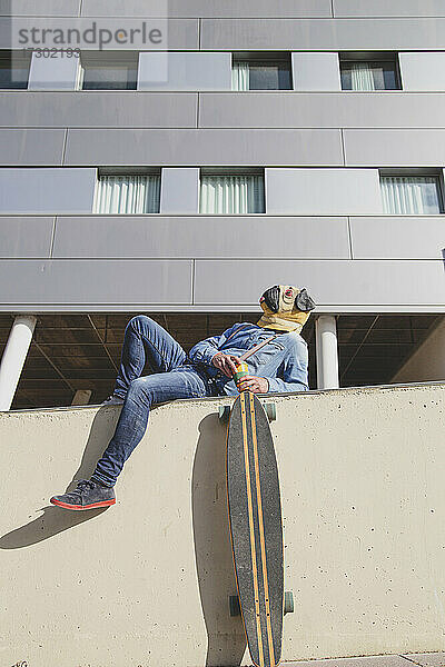 Mann mit Mopsmaske  der auf einer Mauer liegt und ein Longboard trägt.