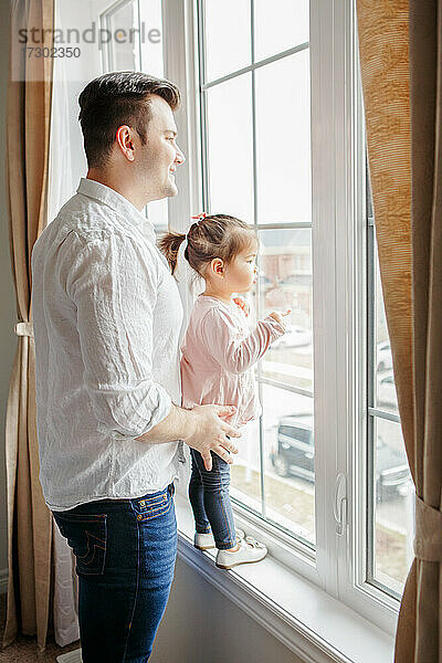 Kaukasischer Vater Vater mit Kleinkind Mädchen Tochter aus dem Fenster schauen