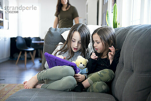 Zwei kleine Mädchen sitzen auf der Couch und benutzen ein Tablet.