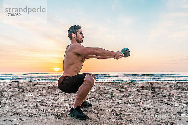 muskulöser Mann trainiert Beine mit Kettlebell am Strand bei Sonnenaufgang