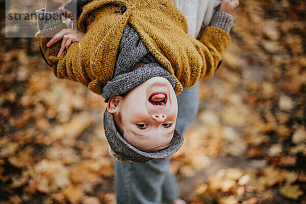 Stilvoller Junge  der sich im Herbst im Stadtpark amüsiert. Glückliches Kind spaziert zwischen gefallenen Blättern. Kindermode. Junge trägt trendigen gelben Mantel  Mütze und Schal. Lächelnder Junge im Freien. Kind springt und läuft