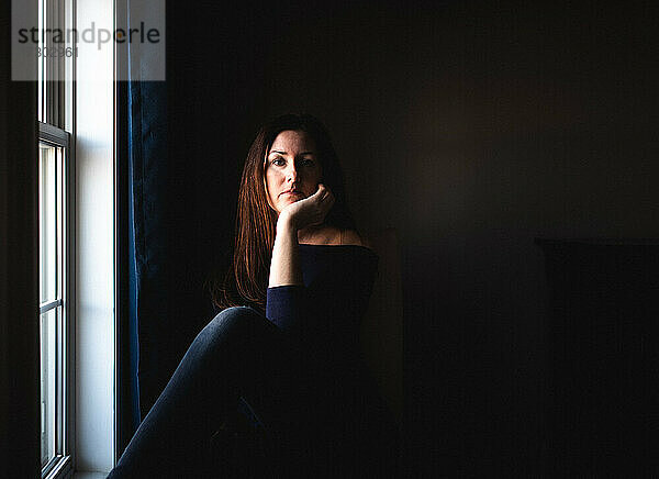 Attraktive Frau sitzt allein in einem dunklen Raum am Fenster.
