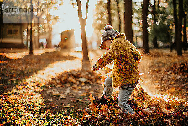 Stilvoller Junge hat Spaß im herbstlichen Stadtpark. Glückliches Kind spaziert zwischen gefallenen Blättern. Kindermode. Junge trägt trendigen gelben Mantel  Mütze und Schal. Lächelnder Junge im Freien. Kind springt und läuft