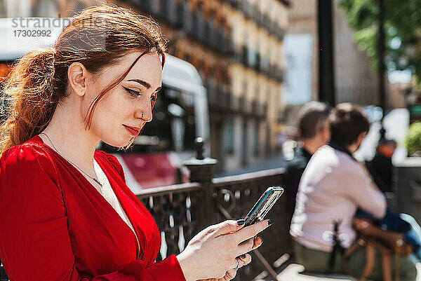 Junges rothaariges Mädchen in roter Bluse benutzt ihr Mobiltelefon auf einem Stadtplatz.
