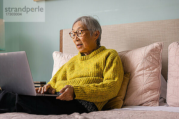 Ältere asiatische Frau  die einen Laptop benutzt  während sie im Schlafzimmer zu Hause sitzt