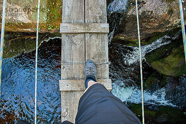 Das Bein eines Sportlers  der eine Holzbrücke mit Seilen über ein natürliches Felsbecken und einen Wasserfall überquert.