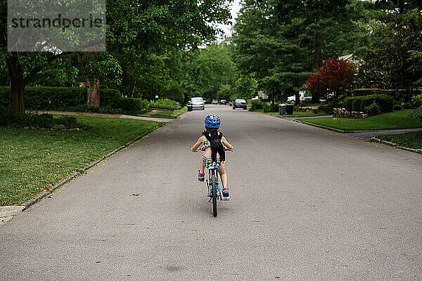 Kleines Mädchen fährt allein auf dem Fahrrad die Straße hinunter  mit einem Spielzeugäffchen auf dem Rücken