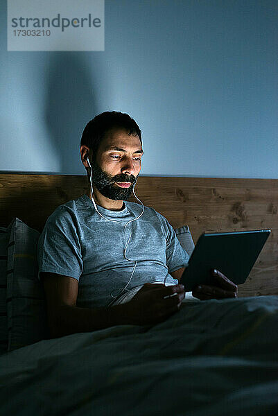 Junger Mann mit Kopfhörern und Tablet auf dem Bett in der Nacht