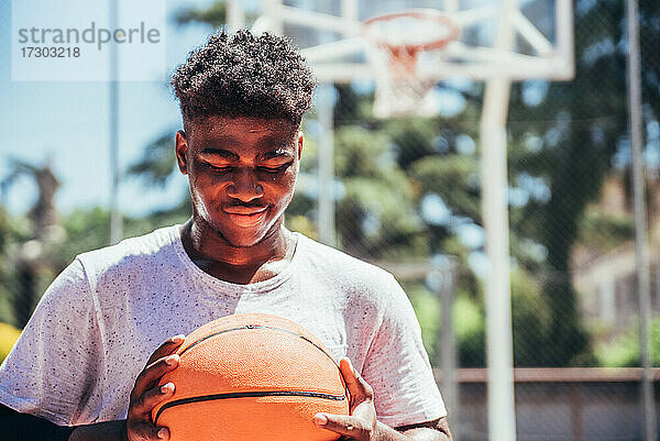 Porträt eines schwarzen afroamerikanischen Jungen  der auf einem städtischen Basketballplatz einen Basketball gegen seine Brust hält.