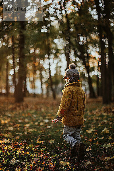 Stilvoller Junge  der sich im Herbst im Stadtpark amüsiert. Glückliches Kind spaziert zwischen gefallenen Blättern. Kindermode. Junge trägt trendigen gelben Mantel  Mütze und Schal. Lächelnder Junge im Freien. Kind springt und läuft