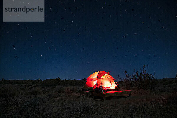 beleuchtetes Zelt auf Campingplattform in der Wüste unter Sternenhimmel