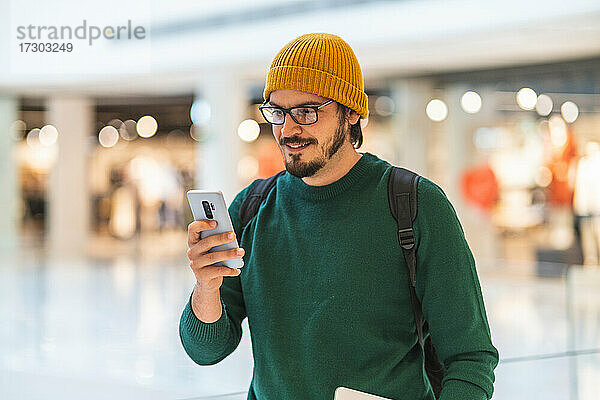 Moderner spanischer Mann lächelt und benutzt sein Smartphone in einem Einkaufszentrum