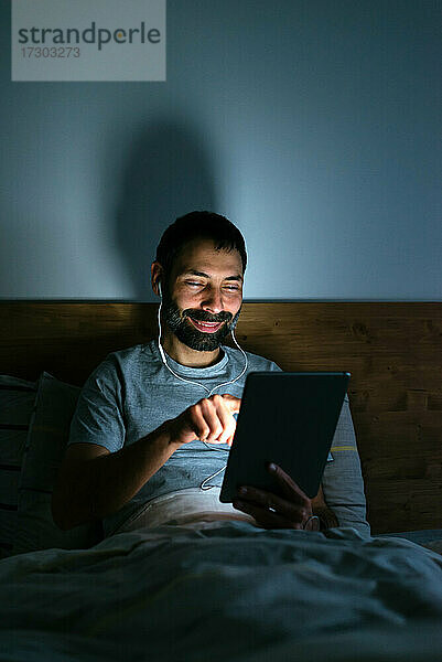 Fröhlicher junger Mann  der nachts auf dem Bett ein Tablet benutzt