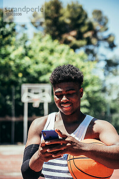 Porträt eines schwarzen afroamerikanischen Jungen  der sein Mobiltelefon auf einem städtischen Basketballplatz benutzt.