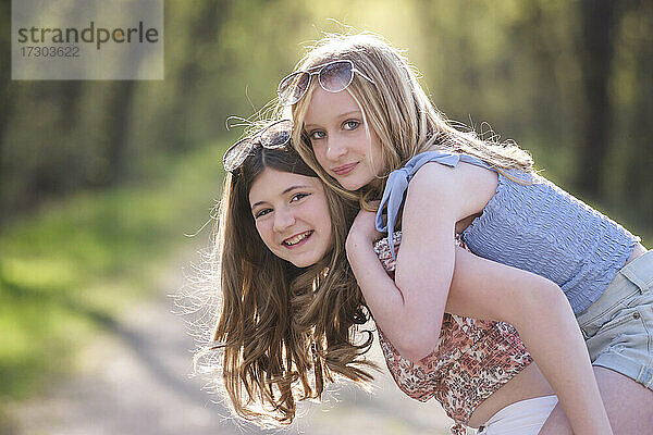 Zwei glückliche  hübsche junge Mädchen auf einer sonnigen  von Bäumen gesäumten Landstraße.