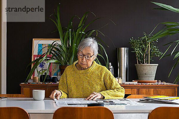 Ältere Frau in gelbem Pullover liest am Esstisch sitzend Zeitung