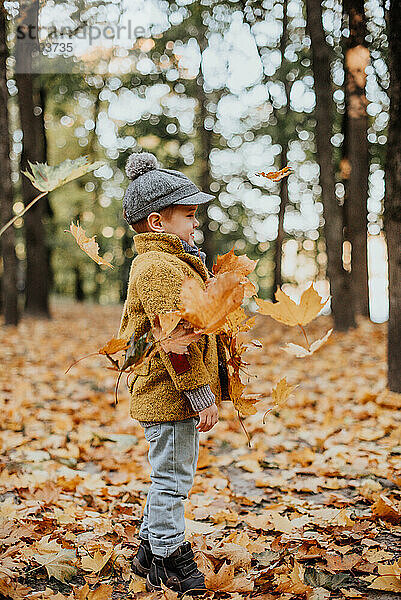 Stilvoller Junge  der im Herbst im Stadtpark Spaß hat. Glückliches Kind spielt mit gefallenen Blättern. Kindermode. Junge trägt trendigen gelben Mantel  Mütze und Schal. Lächelnder kleiner Junge im Freien. Kind springt und rennt