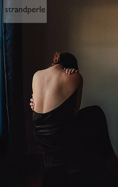 Rückenansicht einer traurigen  nach vorne gebeugten Frau mit entblößtem Rücken in einem dunklen Raum.