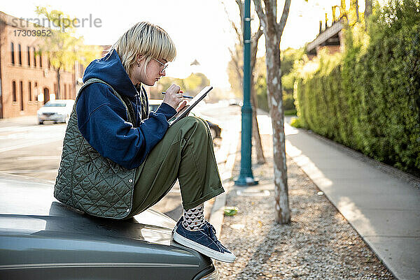 Junger Erwachsener sitzt auf der Motorhaube eines Lastwagens und arbeitet mit Stift und Tablet