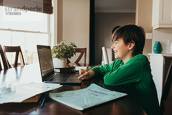 Glücklicher Junge  der am Computer am Küchentisch online Schularbeiten erledigt.