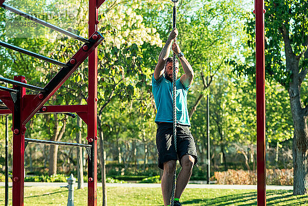 Dunkelhaariger Sportler mit Bart klettert in einem Park auf ein Seil. Outdoor-Fitness-Konzept.