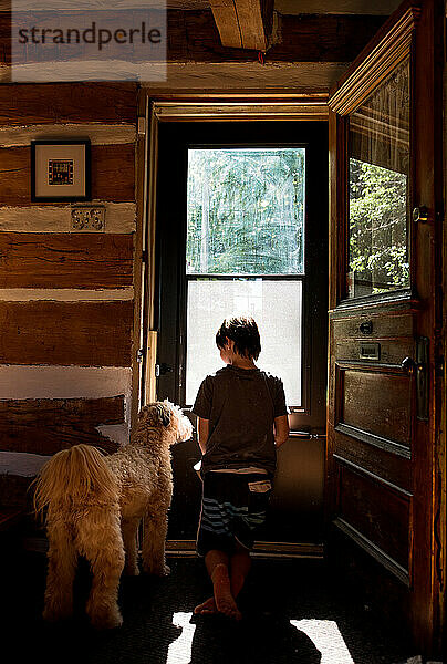 Junge und sein Hund schauen an einem sonnigen Tag aus der Tür eines Blockhauses.