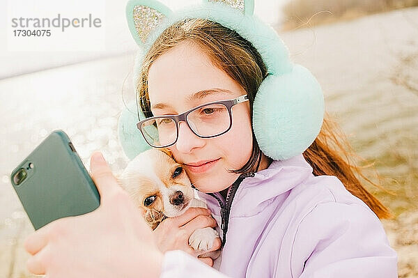 Mädchen macht Selfie auf Smartphone. Porträt eines glücklichen Teenager-Mädchens mit ihrem kleinen Chihuahua-Hund. Ein Mädchen auf dem Hintergrund der Natur umarmt ihren Chihuahua-Hund in der Kleidung.